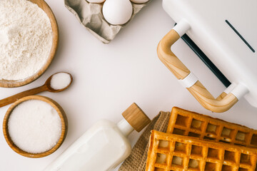 Ingredients for homemade waffles arranged on a white surface, including flour, sugar, eggs, and...