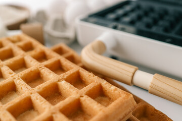 Close-up of a waffle iron with wooden handle, alongside freshly baked waffles and ingredients in a kitchen setting