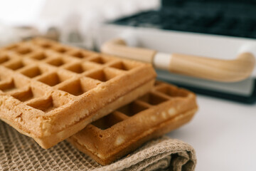 Close-up of a waffle iron with wooden handle, alongside freshly baked waffles and ingredients in a kitchen setting