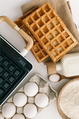 Ingredients for homemade waffles arranged on a white surface, including flour, sugar, eggs, and milk. A waffle maker with wooden handles is visible in the frame
