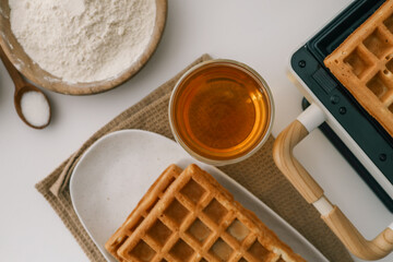 Freshly baked golden waffles served on a white plate with a glass of honey. Crisp texture and inviting aroma. A warm breakfast scene with a waffle maker in the background