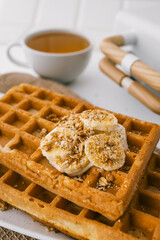 Freshly baked waffles with banana slices, yogurt, and granola on a white plate. A cup of tea and a waffle iron in the blurred background create a warm and inviting breakfast setting