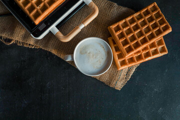 A steaming cup of coffee next to freshly made waffles on a dark textured background, with a waffle maker in the blurred background. Cozy breakfast atmosphere