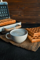 A steaming cup of coffee next to freshly made waffles on a dark textured background, with a waffle maker in the blurred background. Cozy breakfast atmosphere