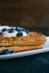 Freshly baked waffles topped with whipped cream, blueberries, and syrup, served on a white plate. A cozy and appetizing breakfast scene with a blurred background