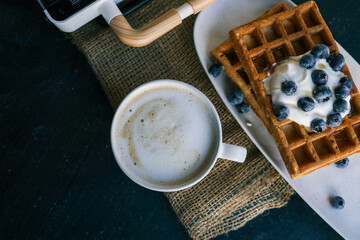 Stack of golden waffles topped with whipped cream and fresh blueberries, served on a rustic plate with a blurred cup of coffee in the background. A cozy, delicious breakfast setting