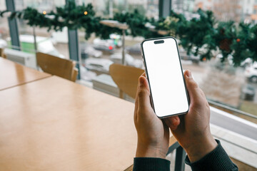 A person holding a smartphone with a blank white screen near a wooden table by a large window. The view outside shows urban buildings and cars with festive decorations on the window