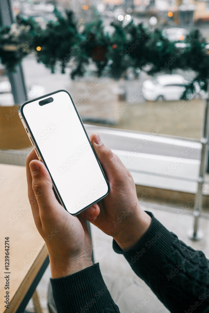 Poster a person holding a smartphone with a blank white screen near a wooden table by a large window. the v
