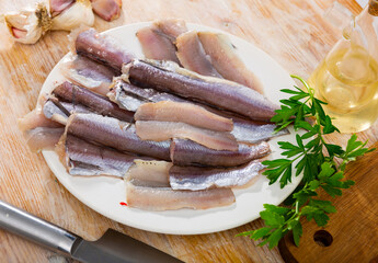 On wooden table there is plate with raw Atlantic fish blue whiting prepared for frying and baking, and necessary spices and ingredients. Process of cutting fish