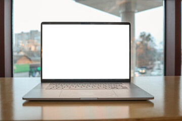 Modern laptop with a blank white screen placed on a wooden table near a large window overlooking a cityscape with buildings. Concept of technology, remote work, and digital workspace