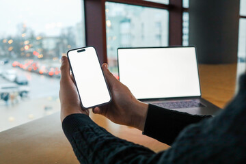 Person holding a smartphone with a blank white screen in front of an open laptop at a wooden desk near a window. Modern workspace, technology, and digital connectivity concept