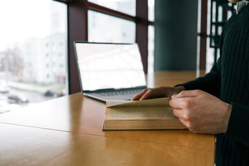 Close-up of hands holding a book and flipping a page, with a laptop in the background on a wooden table near a large window overlooking a blurred urban street scene