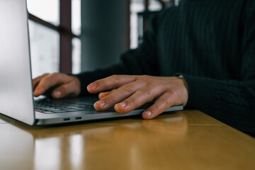 Close-up of a hand typing on a laptop keyboard placed on a wooden table, with large windows providing a blurred urban view in the background, creating a modern workspace atmosphere