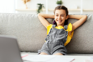 Obraz premium Rest Concept. Smiling female teenager sitting on comfortable couch, leaning back on sofa and looking at laptop screen, watching movie or video, taking break from studying online at home