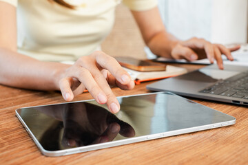 Close-up of a woman's hands using a tablet and laptop on a wooden desk, showcasing modern technology in a home office.