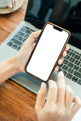 Close-up of a hand holding a smartphone with a blank screen above a laptop on a wooden desk.