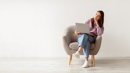Fototapeta premium Deadline stress concept. Frustrated young woman sitting in armchair with laptop computer, looking away deep in thought, feeling tired after long working day against white studio wall, full length