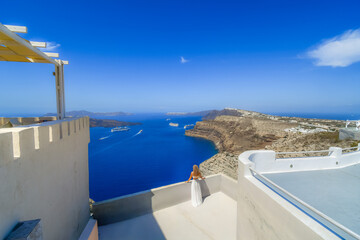 A woman stands on a terrace overlooking the Caldera bay and sea with cruise ships tendered in the...