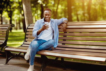 Smiling black man sitting on bench at park and using his smartphone, copy space