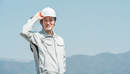青空の下に立つヘルメットを被った作業着姿の男性（建築・土木・建設）
