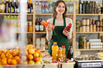 Polite young female seller is kindly offering bottles of natural juice in grocery store
