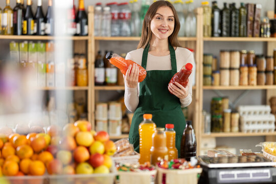Polite young female seller is kindly offering bottles of natural juice in grocery store - Powered by Adobe