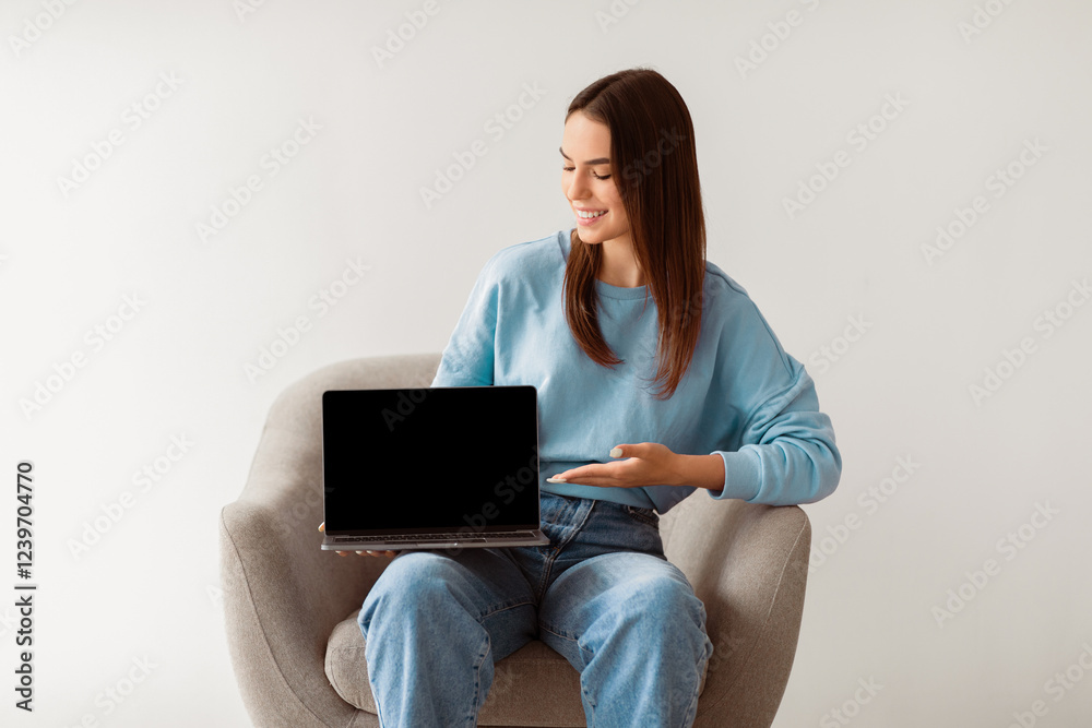 Poster smiling millennial woman in casual clothes showing laptop with empty screen, sitting in armchair on 