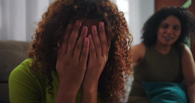 Teenager daughter feeling sad and covering her face with hands, sitting on couch, mother trying to comfort her depressed child in strained family relationship setting