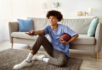 Full length of black teen guy watching American football on TV, holding remote control and ball at home. Cool teenager supporting his favorite rugby team, following sports competition
