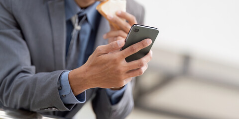 Close up of a young Asian businessman holding a smartphone and a snack in a modern office environment.