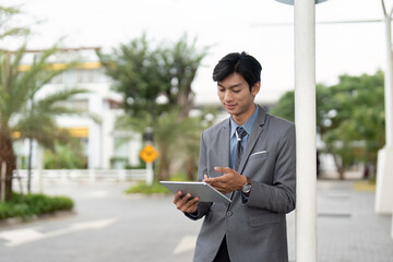 Young Asian business professional smiling while using a tablet outdoors, demonstrating engagement and modern technology in a professional setting.