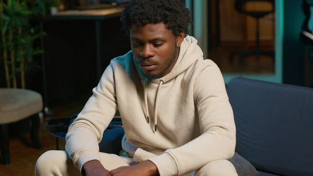 African american male remote worker sitting at home on couch late at night, overthinking about problems. Young man struggling to finish work project before deadline, finding solutions. Camera B.