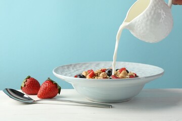Pouring milk into bowl with oatmeal, berries and hazelnuts at white wooden table against light blue background, closeup