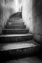 Weathered stone steps wind upward through a narrow, aged stone passage, creating a dramatic grayscale image.