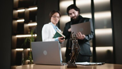 Two business professionals in a courtroom or office, reviewing data or consulting with a laptop, next to a table and the statue of Lady Justice. Concept: professional legal consultations.