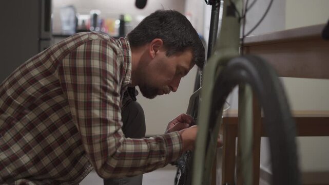 Man is fixing bicycle at home. Male repairs his bike at home. Guy is adjusting his cycle at their home. Setting up a bicycle in a small apartment. Making settings for a cycle in a little studio room