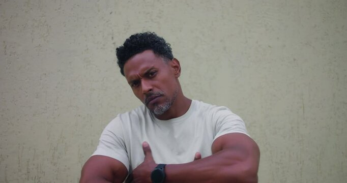 Man of African descent standing against textured wall with arms crossed, intense stare at viewer, tough attitude, strong presence, confrontational expression, dominant posture