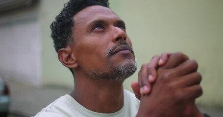 Man of African descent looking up to the sky, hands clasped in deep prayer, hopeful expression, seeking guidance and faith, spiritual connection, urban background, peaceful moment
