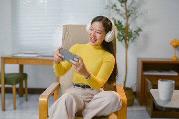 Young woman playing mobile game while wearing headphones and sitting comfortably in armchair at home