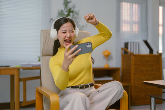 Excited young woman celebrating online game victory using smartphone and headphones at home