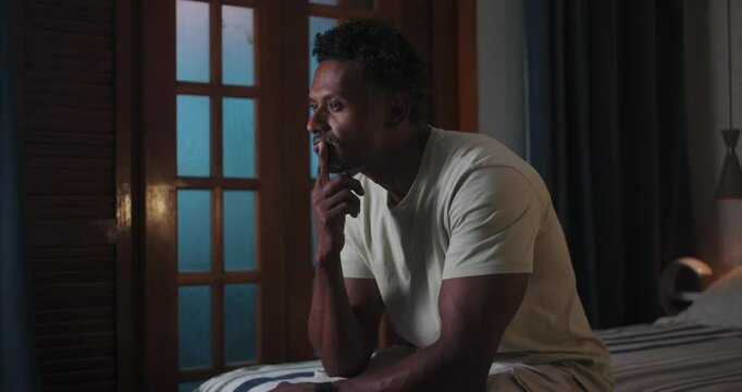 One Black man sitting on bed, leaning forward with hand on chin, deep in thought, introspective mood, serious expression, quiet indoor setting with wooden shutters