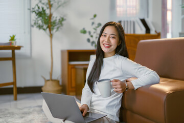 Young asian woman working from home using laptop and drinking coffee
