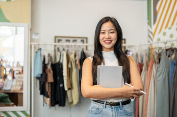 A confident, beautiful Asian female clothing store stands in her store with a tablet in her hand.