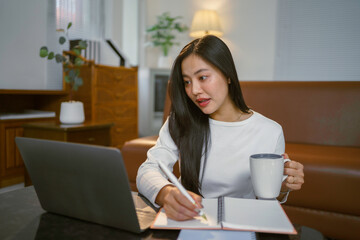 Young Asian Woman Working From Home and Taking Notes While Drinking Coffee