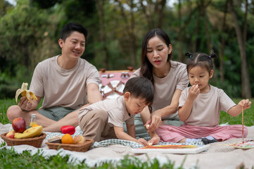 A cheerful Asian family enjoys a picnic in a green public park, sitting on a blanket, and eating.
