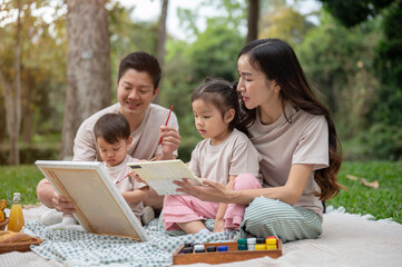 Lovely Asian parents take their kids on a picnic in a park, enjoying painting on a canvas together.