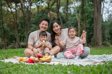 A happy Asian family sits on a picnic blanket in a green park, enjoying their weekend together.