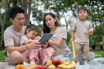 A happy Asian parents are taking their little kids on a picnic in a green park.