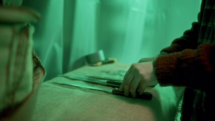 Horror movie scene of torturer placing sharp tools, including knives and saw, laid out on fabric-covered table in room illuminated by dim green light. Close-up, tilt-up shot