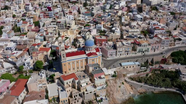 Experience a breathtaking aerial point-of-view shot of the Church of Agios Nikolaos at Asteria Beach, Syros Island, Greece. Marvel at its bright red roof and blue dome set within a cinematic cityscape